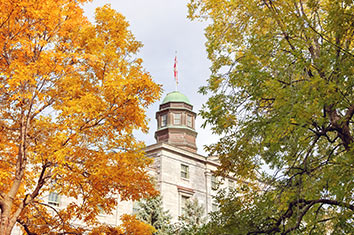 A tower on a brick building, visible between two trees, one with orange leaves and the other with green leaves.