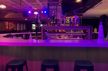 A bar with a white countertop is bathed in blue and purple light. In front of it are three black, metal bar stools.