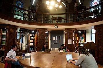Four people sit at a large table. Three of them are using laptops. They are in a circular room surrounded by book shelves, a balcony, and stained-glass windows.