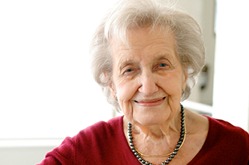 A woman with short white hair, wearing a red shirt and two necklaces, smiles at the camera.