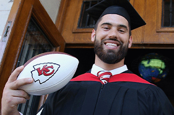 A bearded man in a black and red graduation robe and cap looks down, smiling, into the camera while holding a football labelled KC.
