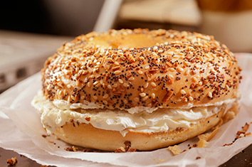 A toasted bagel with sesame and poppy seeds. Cream cheese is spread on both halves of the bagel, which sits, closed, atop a white paper bag.