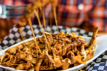 A fork pulls a cheesy bite from a bowl of poutine (French fries covered in cheese and gravy) atop a piece of black and white checkered paper.