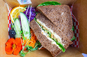 The inside of a brown cardboard box, seen from above, containing two halves of a sandwich on pumpernickel bread next to a salad (cucumber wedges, sprouts, a blackberry, an orange wedge, grated carrot, and an orange nasturtium flower).