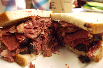 A smoked meat sandwich on untoasted white rye bread, halved, on a white plate. A takeout coffee cup is visible in the background.