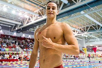 Un nageur, Clément Secchi, se tient debout, la main levée près de la poitrine, dans un stade de natation bondé.