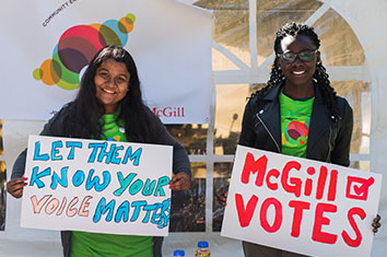 Deux étudiantes de McGill font face à la caméra avec de grandes affiches sur lesquelles on peut lire LET THEM KNOW YOUR VOICE MATTERS et McGill VOTES .