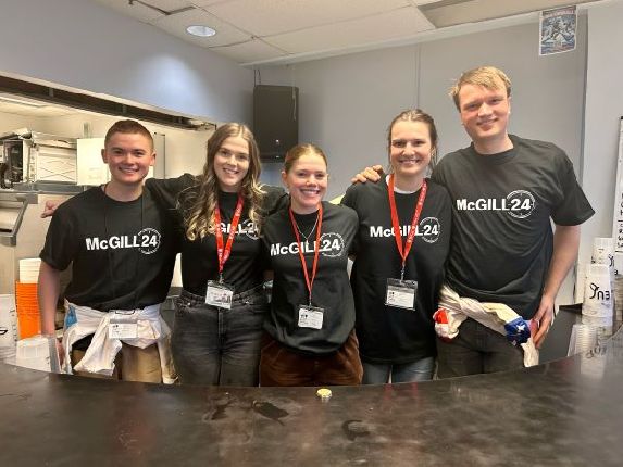 A group of 5 people behind a counter looking at the camera and smiling. Wearing black t-shirts that say McGill24