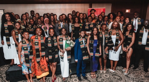 A large group of people in formal attire wearing matching patterned stoles stands together in several rows at an indoor event, with one person near the center holding a bouquet of flowers.