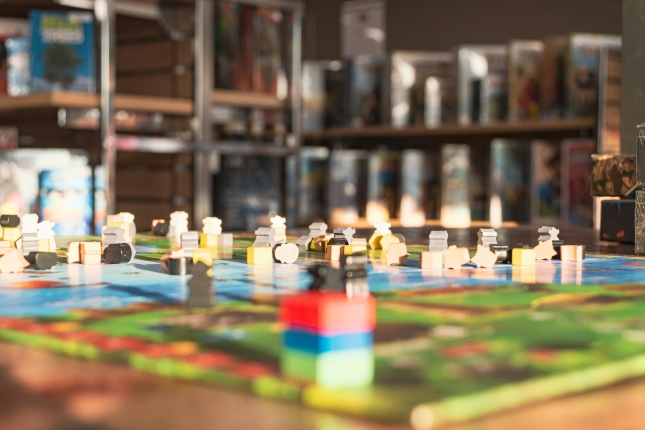 Colorful wooden board game pieces spread across a game board on a table, with shelves of boxed games in the background.