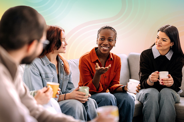 Four young people sitting around a couch each holding a cup of coffee and talking