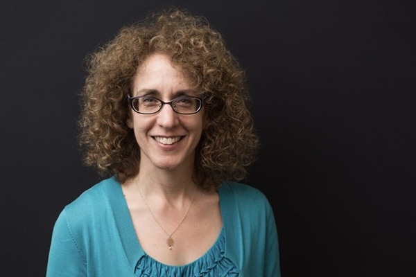 Professional headshot of a person with curly hair and glasses wearing a teal top and cardigan, posed against a dark studio background.
